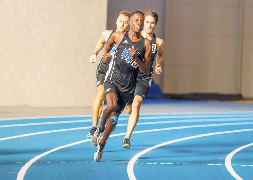 Three male runners compete on an indoor blue track, with the foreground athlete wearing a black singlet marked “GV L” sprinting around the curve. Two competitors trail close behind as the lead runner pushes forward with focused determination.